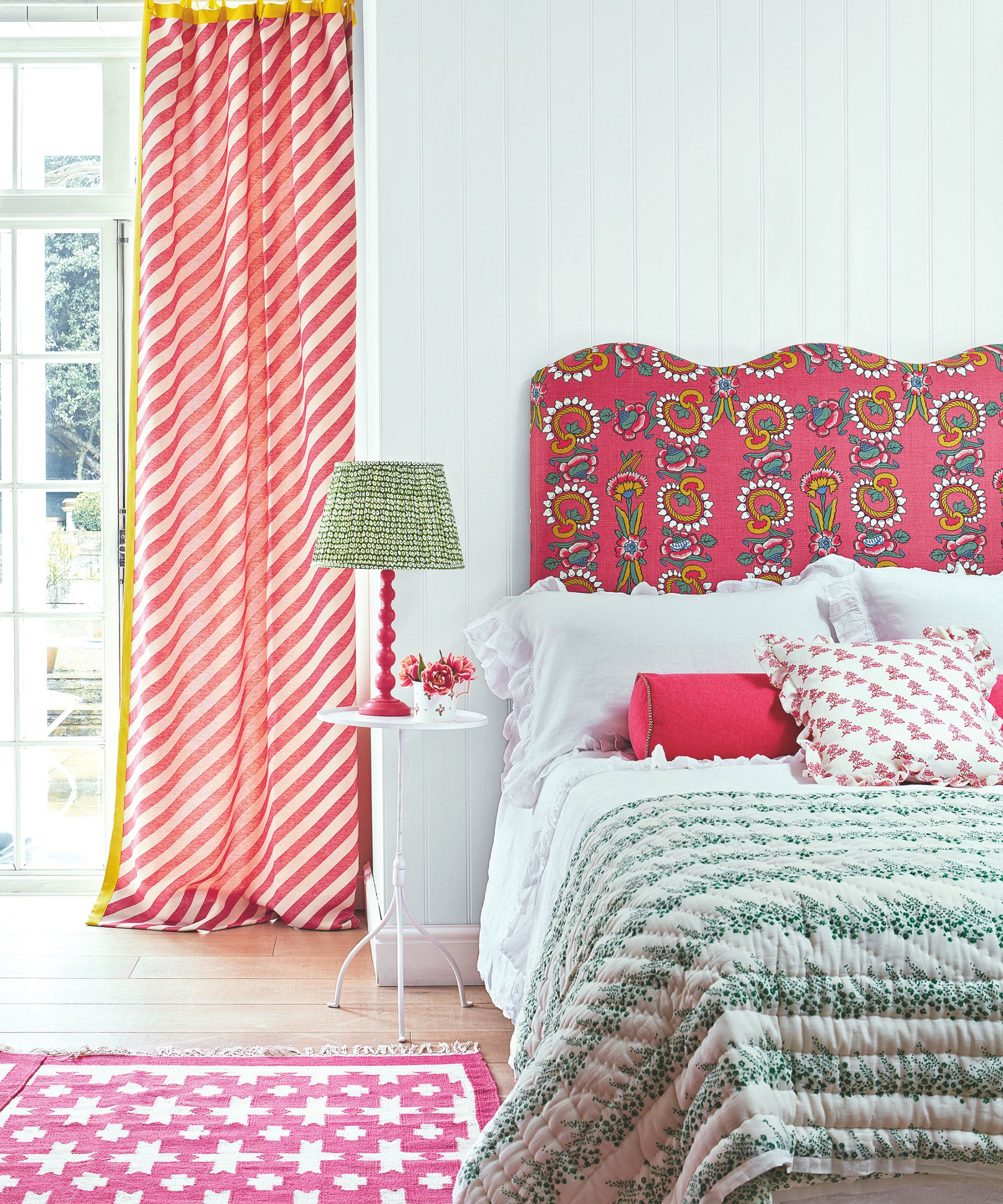A colorful bedroom with white panelled walls, and a pink patterned headboard. The bedding is white, with pink cushions and a green bedspread, and a pink patterned rug to the left, on wooden flooring. To the left is a large mirror with red and white striped curtains.