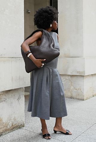 a photo showing the best summer-to-fall transition items with a woman standing against columns wearing a gray top, gray pleated skirt, black mules, and a large brown woven leather handbag