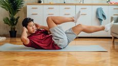 A man performs a bicycle crunch on an exercise mat at home. He is on the floor, with his lower back and buttocks in contact with the mat, while his neck, head and legs are elevated. His hands are behind his head and he is twisting to the side so that his right elbow stretches to meet his bent left knee. Behind him we see drawers and a plant.