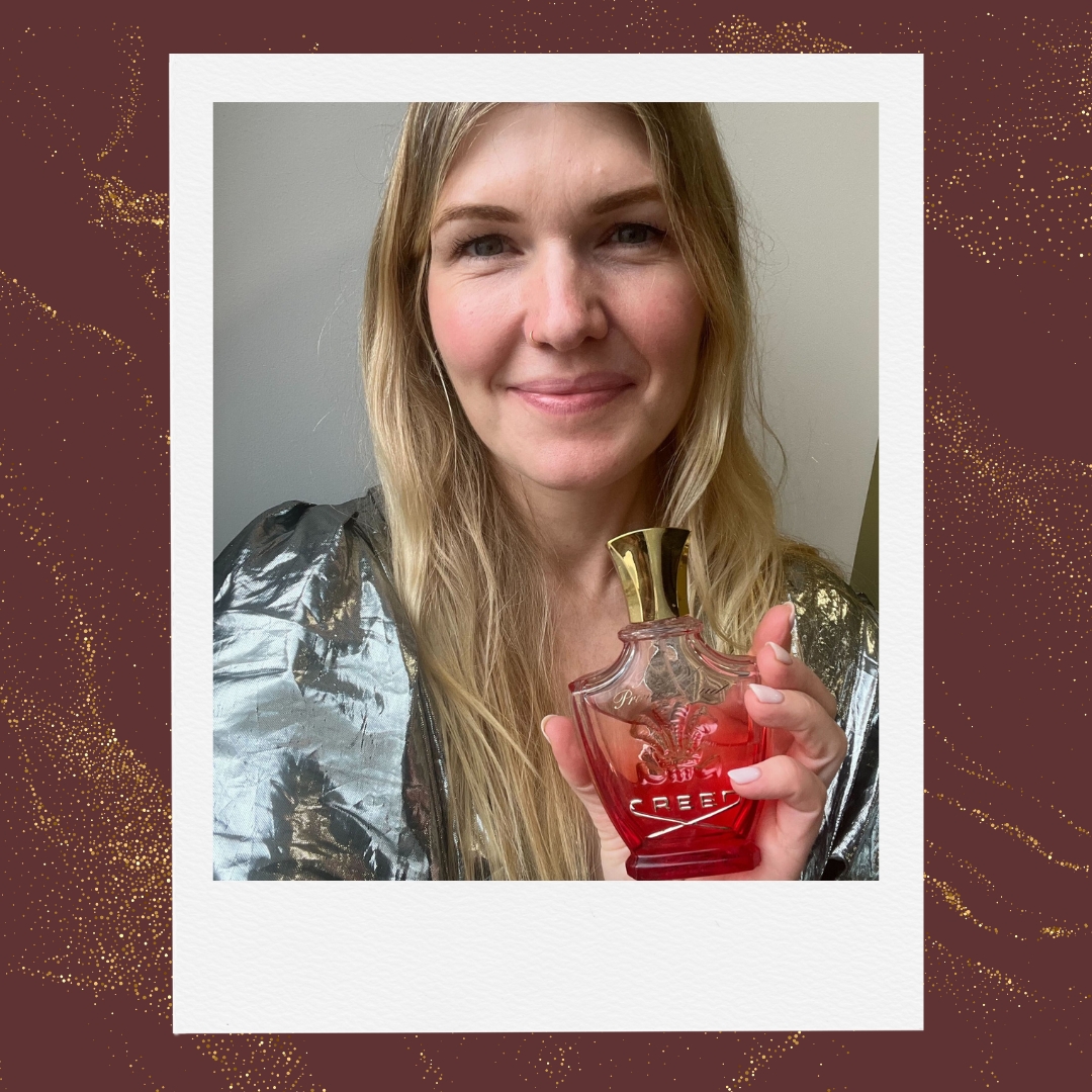 A picture of digital beauty channel editor, Fiona McKim, seen wearing a silver shiny top and holding a bottle of Creed Royal Princess Oud perfume/ featured in a burgundy background with a gold glitter overlay