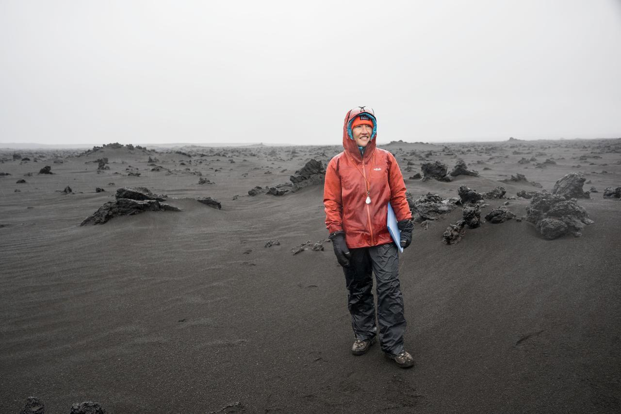 A woman in a bright orange jacket and brown pants stands in a muddy dark landscape