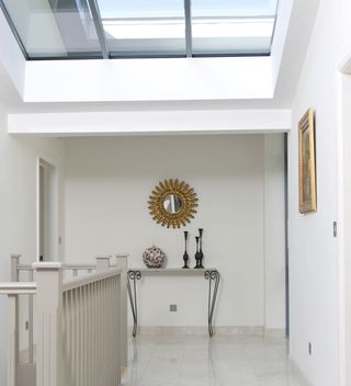 A white, light and airy landing with a skylight above, a small console table at the end underneath a gold sun mirror