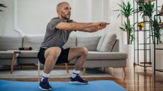 A man in shorts, t-shirt and sneakers executes a squat in a living room on a yoga mat. His knees are bent, thighs parallel with the floor and arms straight out in front of his shoulders. Behind him is a sofa, tall plants and a coffee table.