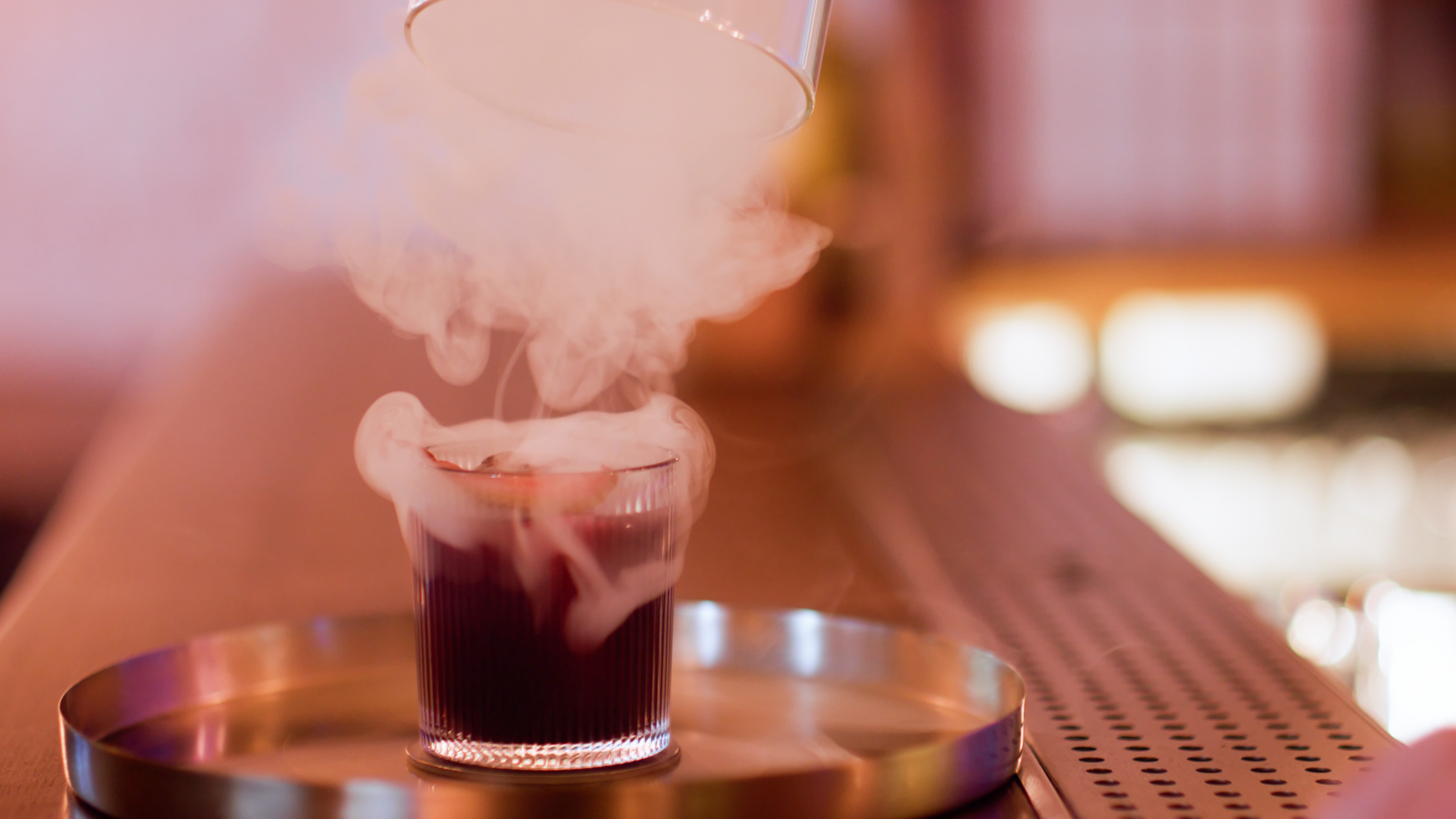 a cloche is being lifted from a cocktail sitting on a bar, releasing a cloud of white smoke 