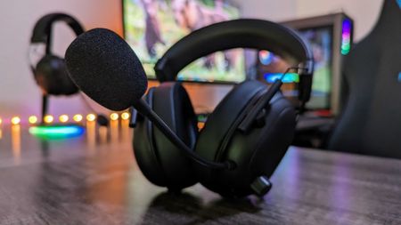 A black gaming headset with a microphone is on a desk in front of a colorful, blurred gaming setup. The ambiance is vibrant and tech-focused.
