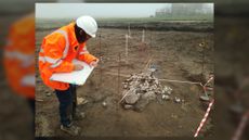 We see a woman in an orange jacket doing an archaeological survey near a dirt field.