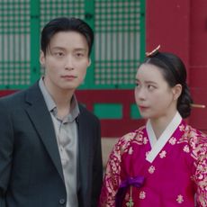 A Korean man in a suit (Heo Nam-jun as Cha Se-gye) and a Korean woman in traditional clothing (Lim Ji-yeon as Shin Seo-ri) stand in front of a traditional palace in 'My Royal Nemesis'.