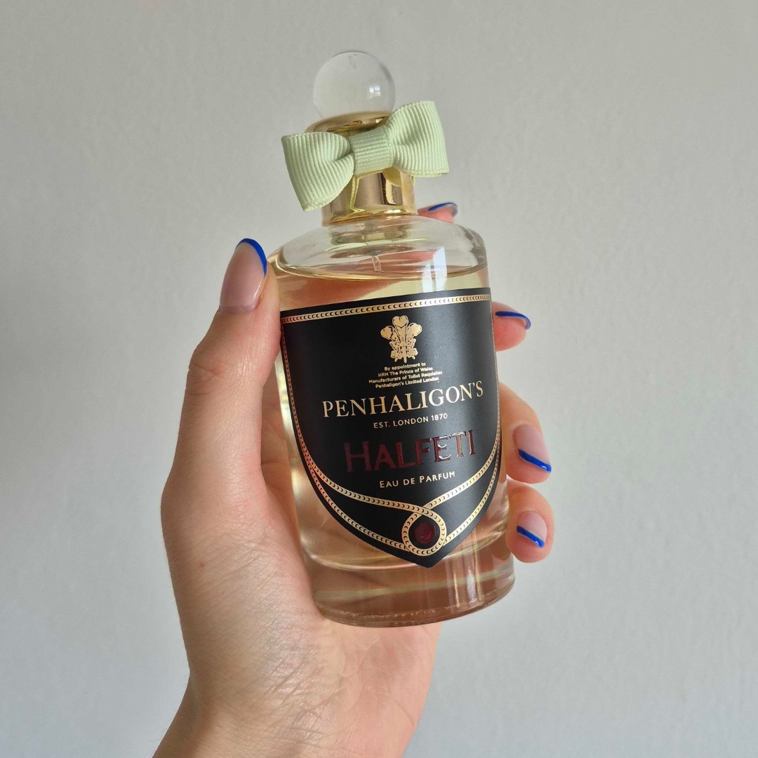A close-up of Digital Beauty Writer Naomi Jamieson's hand, seen holding a bottle of Penhaligon's Halfeti perfume, which features a green bow/ in front of a cream wall background