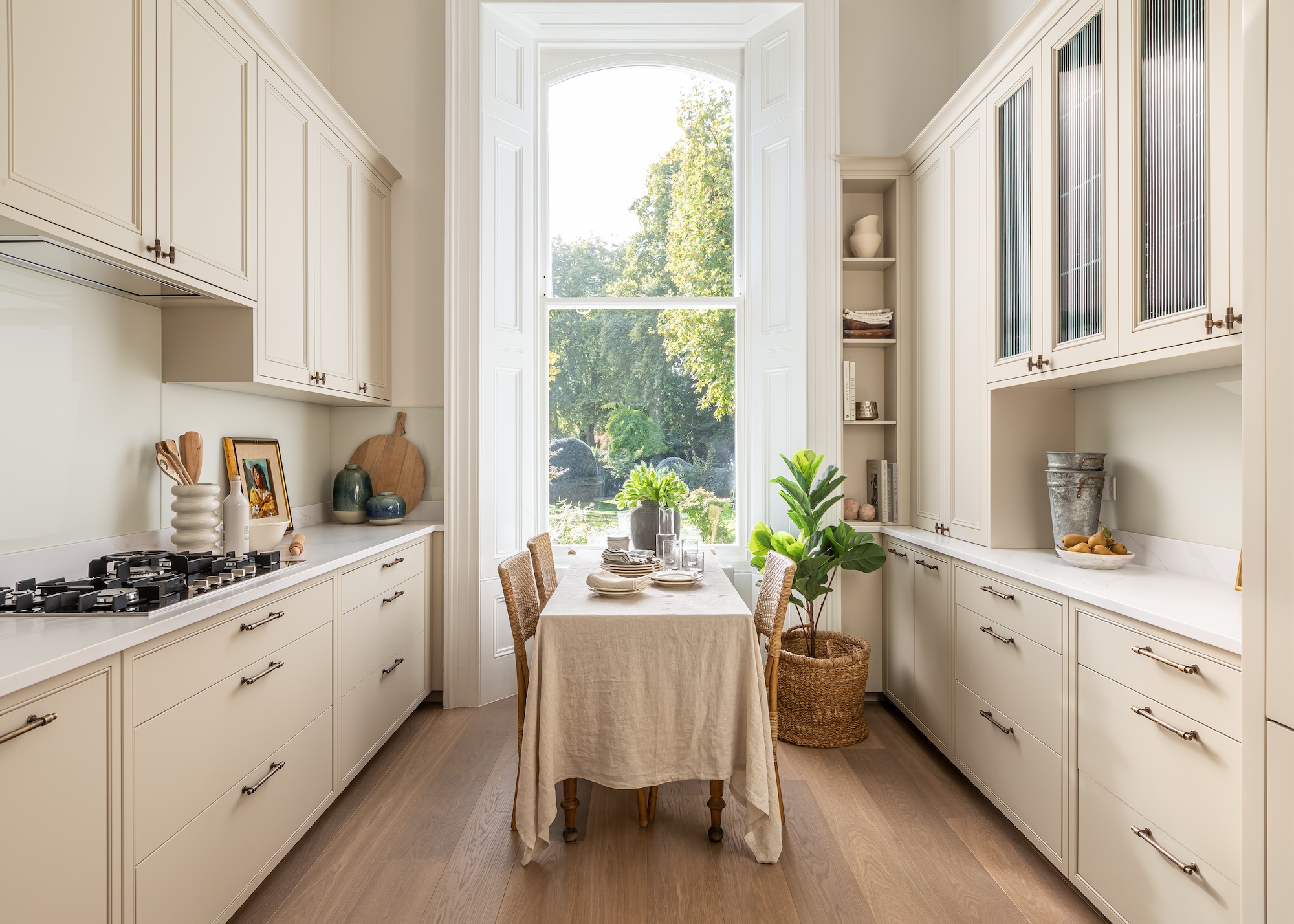 A cream colored kitchen with lower and upper cabinets. There are three glass fronted cabinets on the right and a view of a slender open shelving with accessories and books on it.