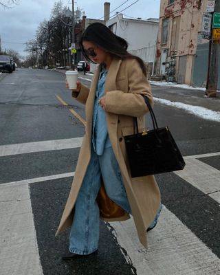 Sasha Mei wearing a camel coat, denim shirt, and jeans with a black tote bag.