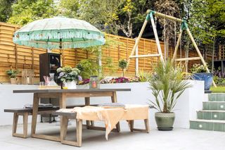 Patio with light grey tiles, wooden dining bench set, green parasol and tiled steps to lawn