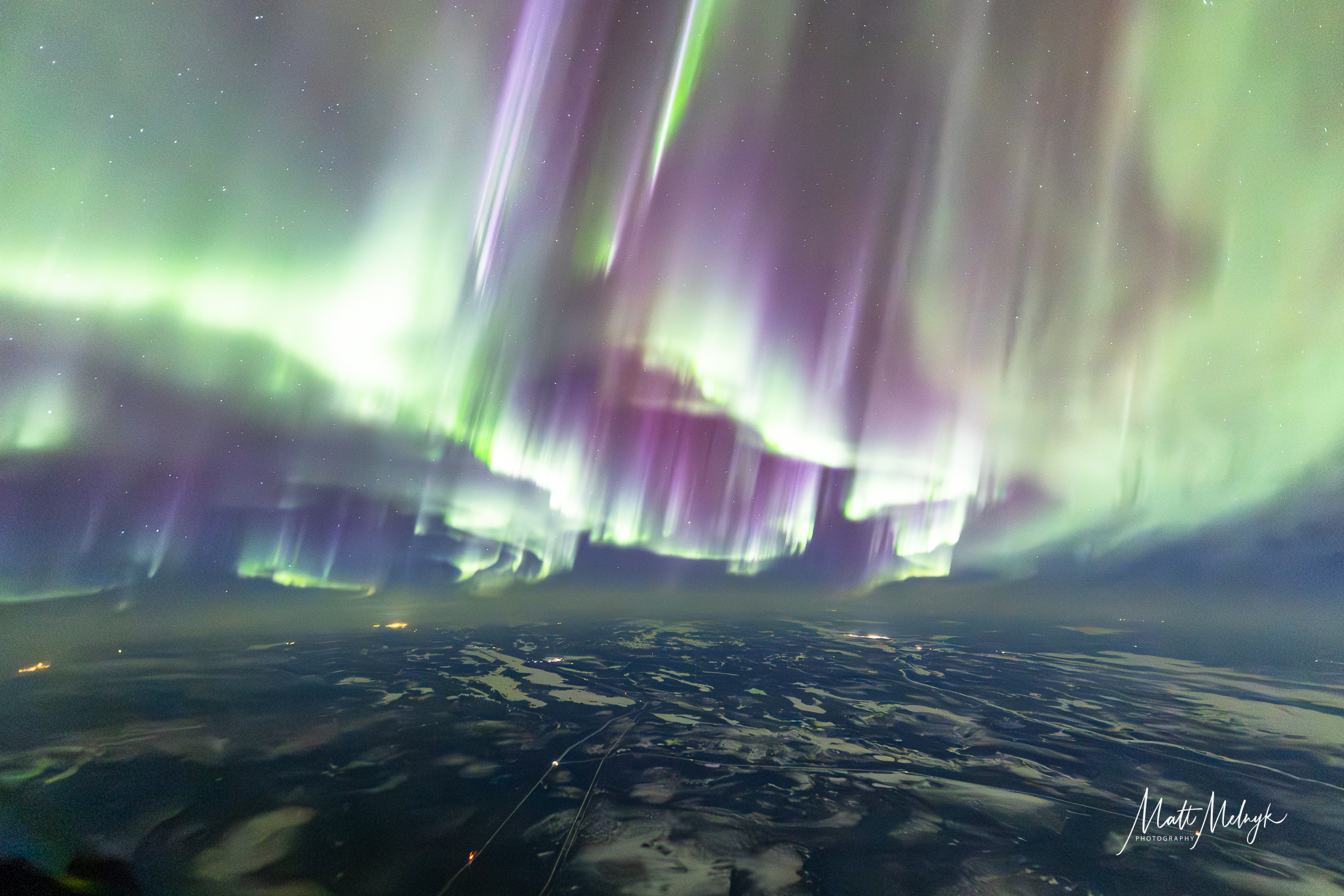 curtains of green auroras fill the sky, with broken clouds below and distant glows of city lights.