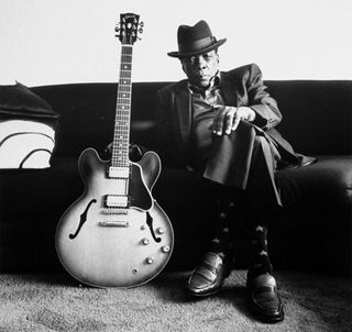 Blues singer John Lee Hooker wearing hat, posing on couch w. his guitar standing on the floor next to him at home, 07 October, 1990