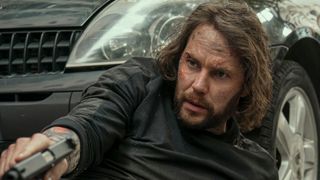 An injured man sits in front of a car while pointing a gun at something offscreen