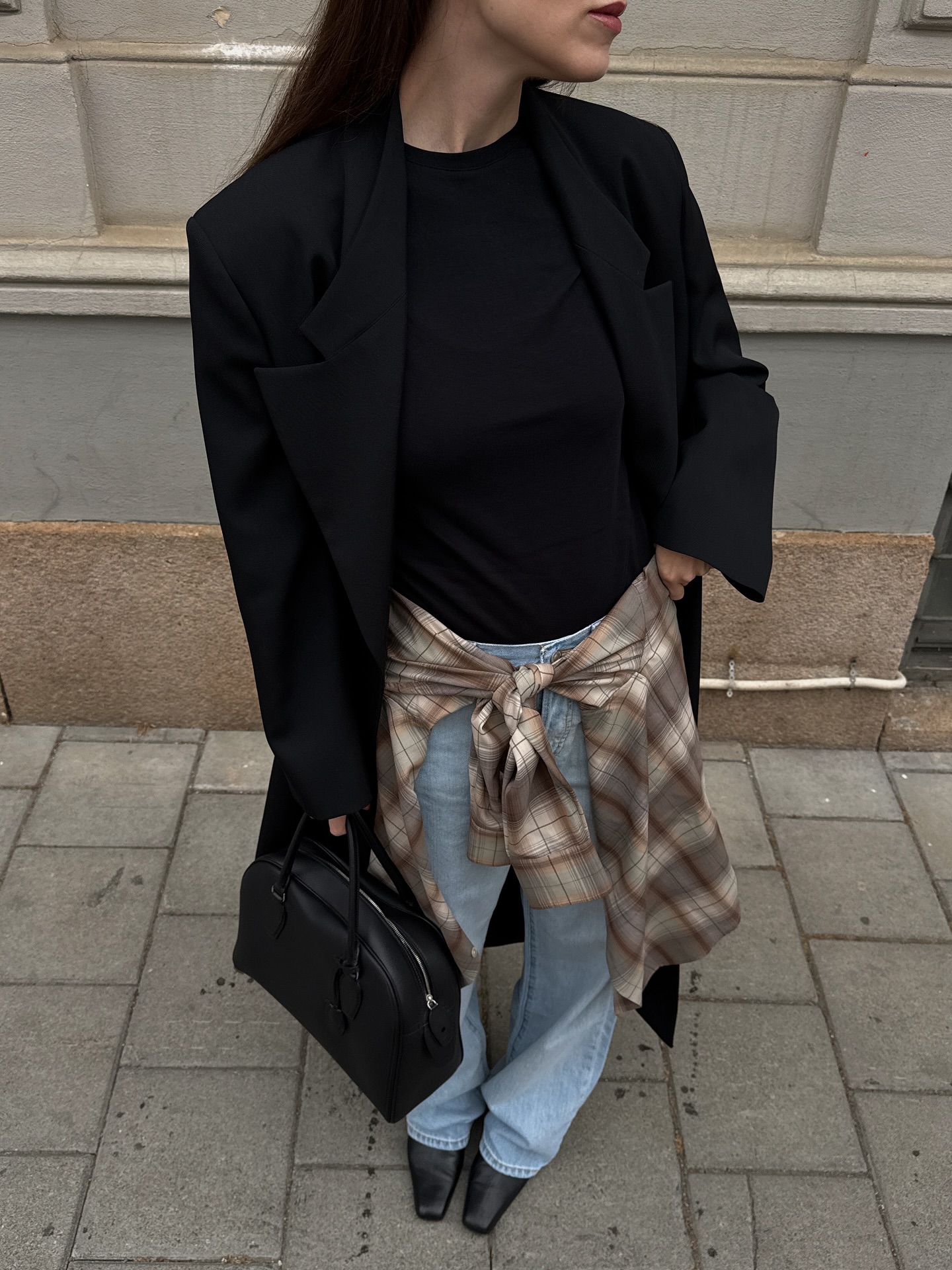 Woman wears black coat, black t-shirt, checked shirt, blue jeans, black boots, carries black The Row India bag