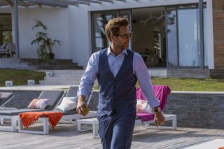 Harry King (Jamie Bamber) walks in the back garden of a French villa, dressed in a smart waistcoat and trousers, with sun loungers behind him