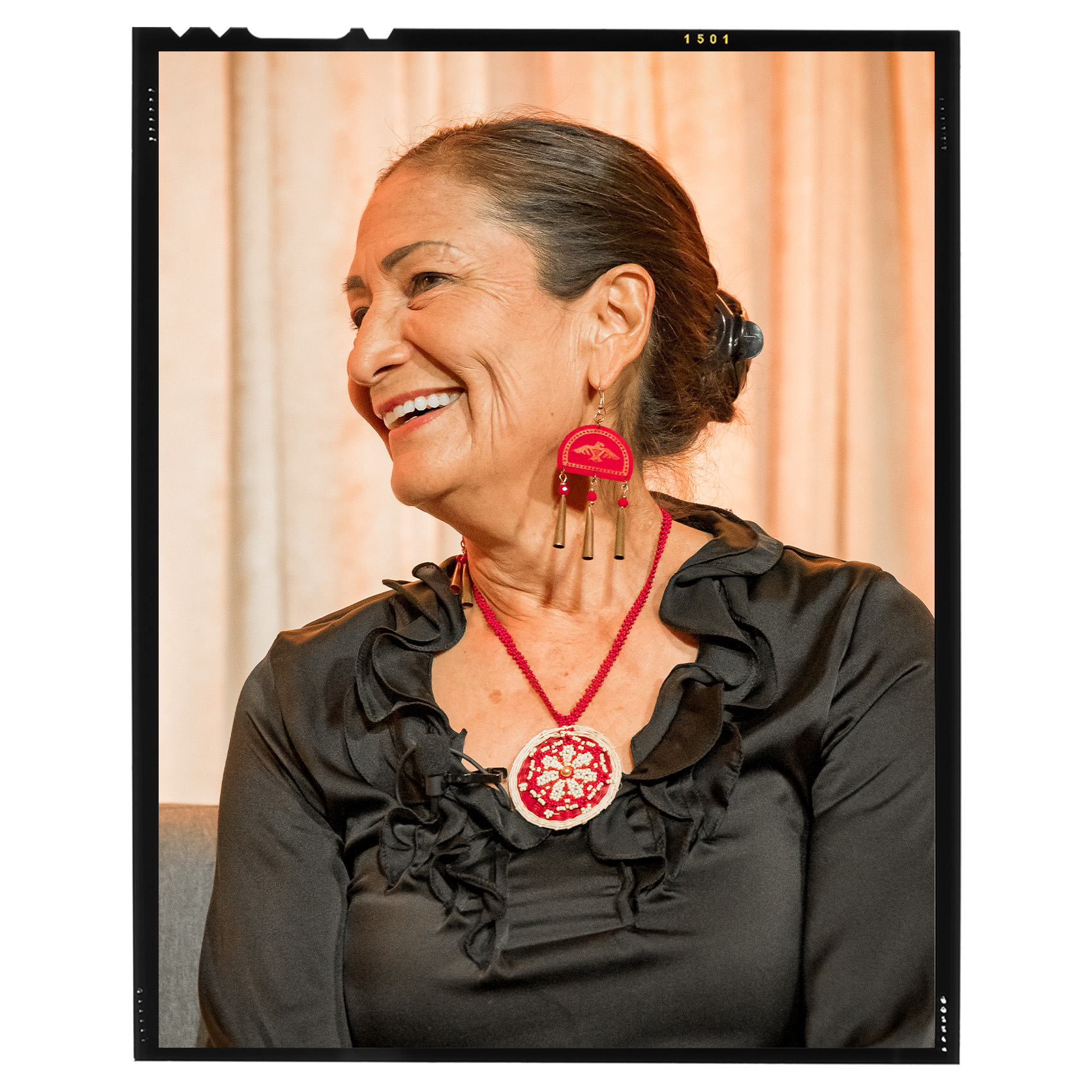 portrait of Deb Haaland in a gray ruffle dress, red and white beaded necklace, and red and brown chandelier earrings