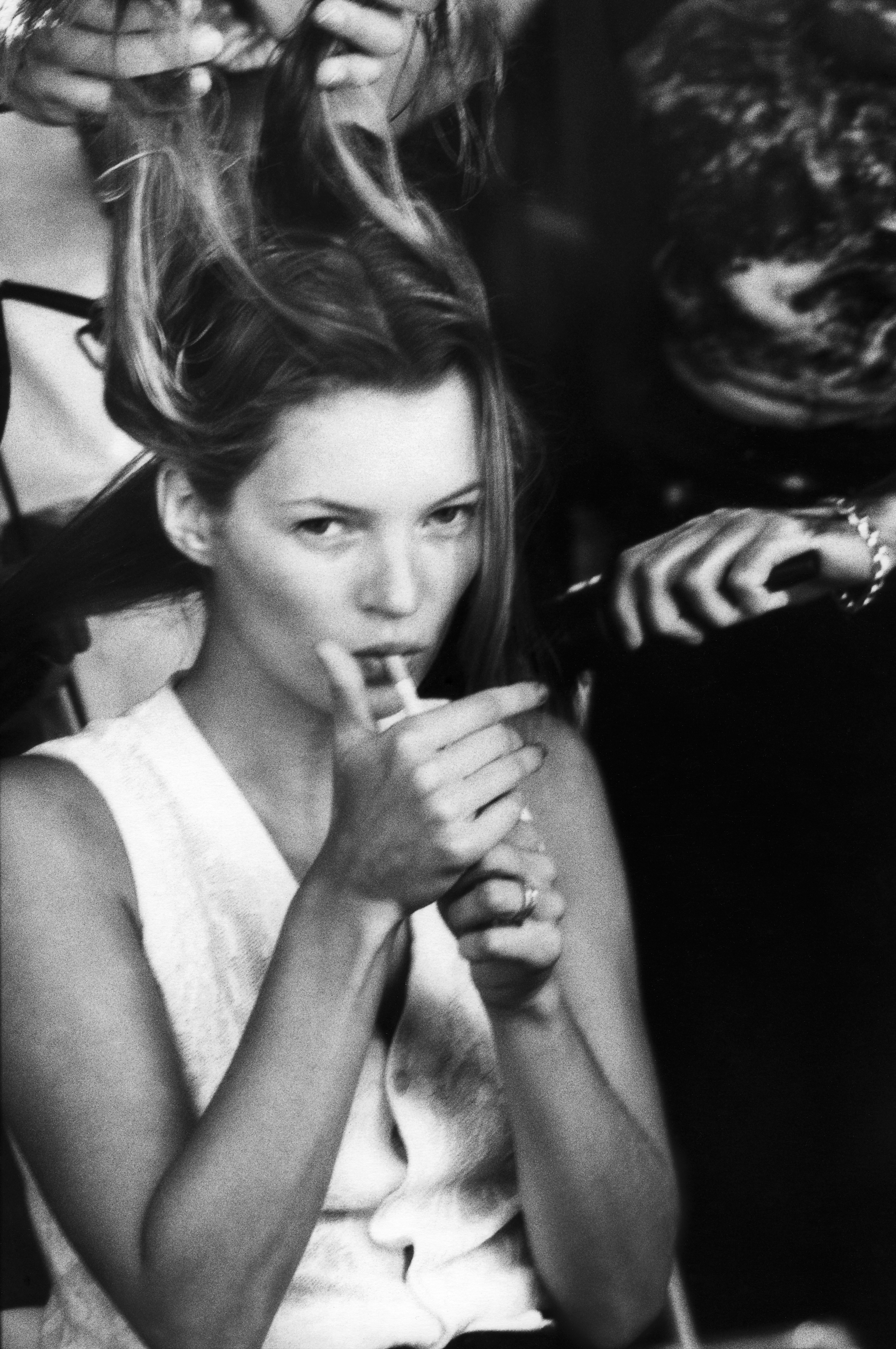 Kate Moss lights a cigarette in a crowd, her hair caught mid-movement, in a candid black and white photograph that has the energy of an unguarded moment rather than a posed portrait.