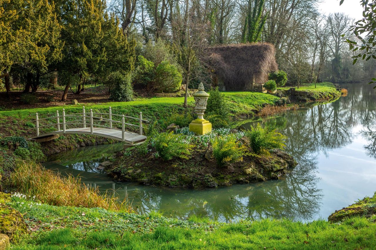 An ornamental bridge leads to a rockwork island in the lake. Behind is the twiggy-roofed boathouse. Eythrope Park snowdrops. ©Clive Nichols