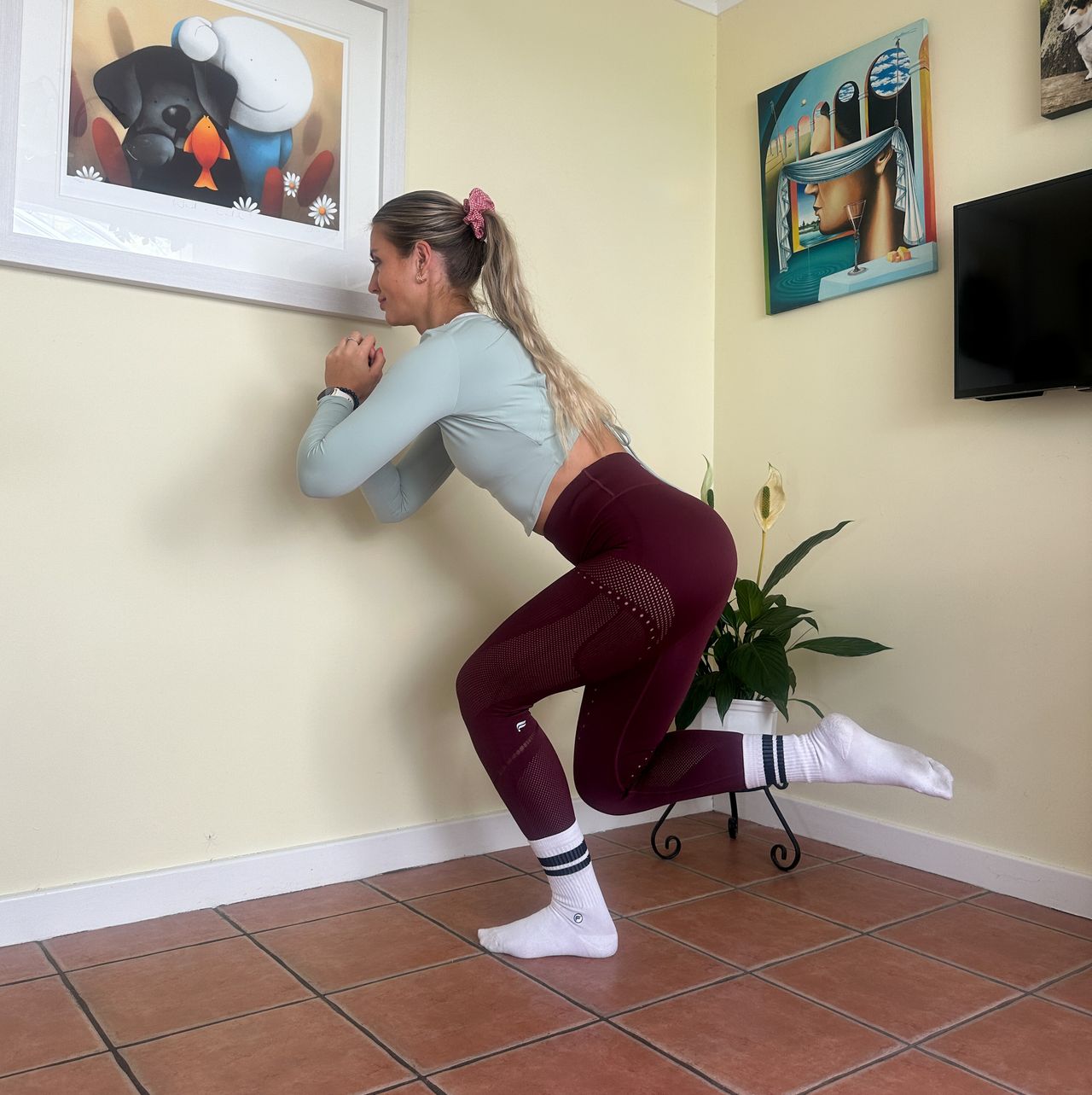NASM qualified trainer Chloe Thomas performs a counterbalance squat in a room with cream colored walls and pictures on the walls. She is standing on one leg, leaning forward slightly and squatting down.