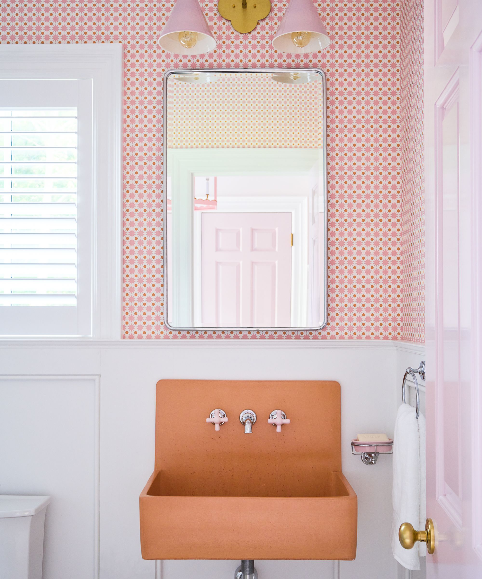 A small bathroom with pink and orange small-scale patterned wallpaper, white panelling, an orange sink, and pink wall sconces.
