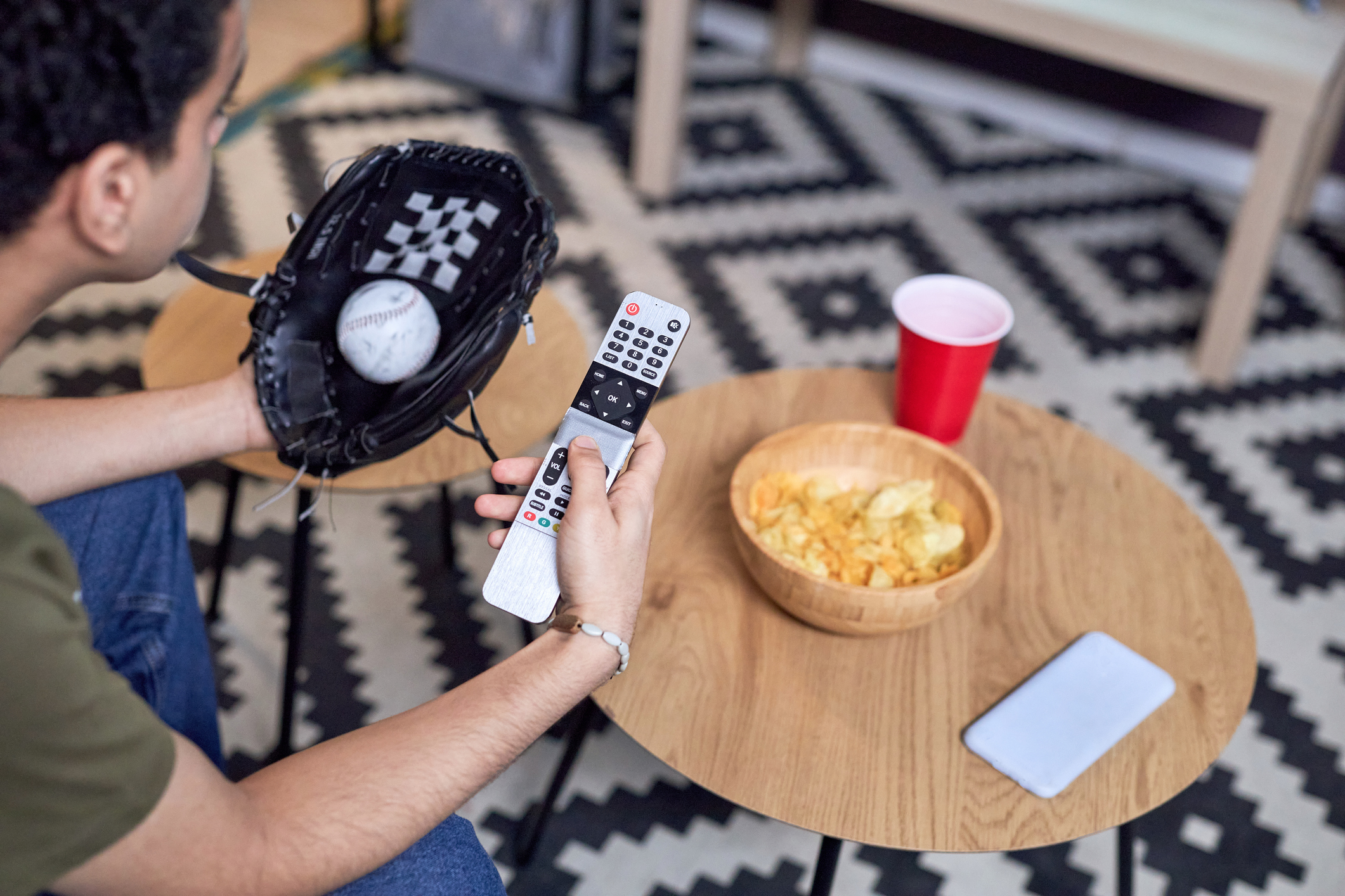 High angle closeup of baseball fan holding TV remote with phone and snacks on table in front of him