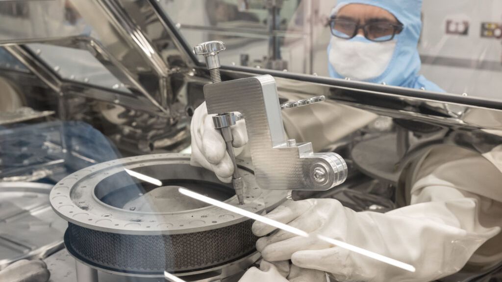 A scientist in clean room suit attaches tool in glove box on asteroid sample capsule