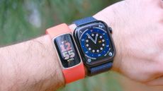 Close up of a Fitbit Charge 6 with an orange band on a user's wrist next to an Apple Watch 10 with an out-of-focus green background