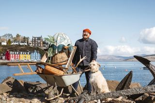 Designing The Hebrides season 2: Banjo Beale stands on a harbour wall with the brightly coloured houses of Tobermory visible in the distance behind him. He is clutching a wheelbarrow piled high with chairs, picture frames, lampshades and so on. Next to him is his dog, Grampa.