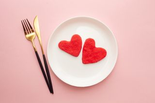 Red heart-shaped cookies on a white plate with a gold knife and fork, on a light-pink background.