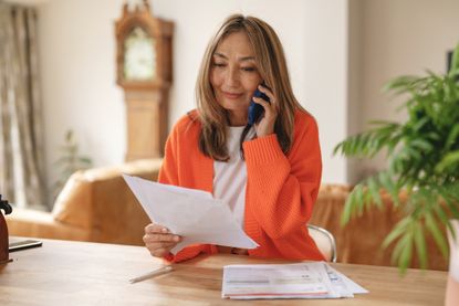 A woman sits on a chair in a living room, talking on a phone and holding papers in her hands.