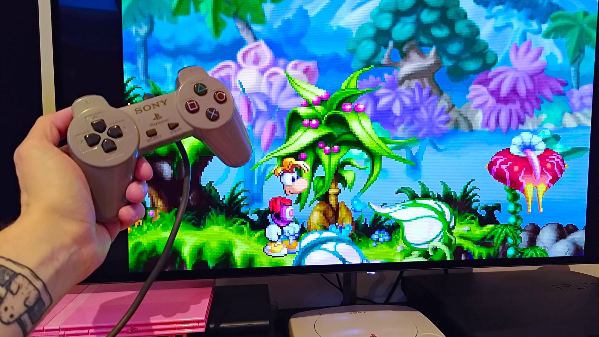 Hand holding PS1 controller next to Rayman gameplay on monitor.