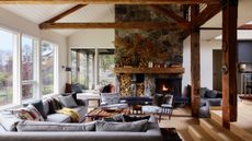 Rustic living room with exposed wooden ceiling beams, a large stone fireplace with built-in log storage, and comfortable gray sectional seating around a coffee table.