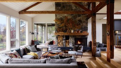 Rustic living room with exposed wooden ceiling beams, a large stone fireplace with built-in log storage, and comfortable gray sectional seating around a coffee table.
