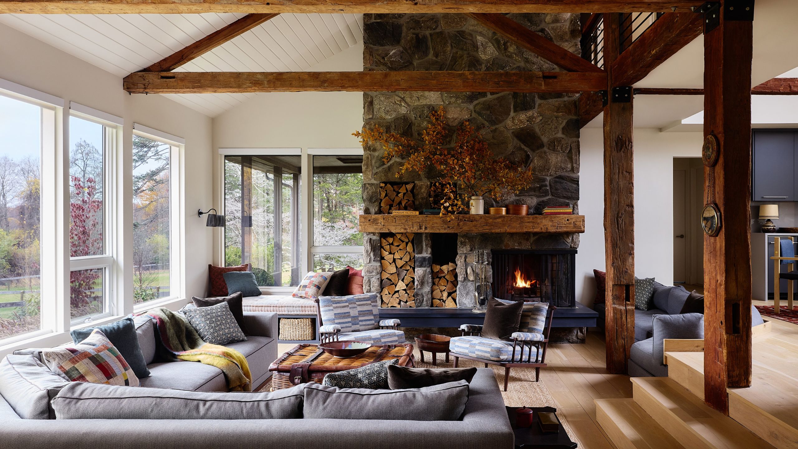 Rustic living room with exposed wooden ceiling beams, a large stone fireplace with built-in log storage, and comfortable gray sectional seating around a coffee table.