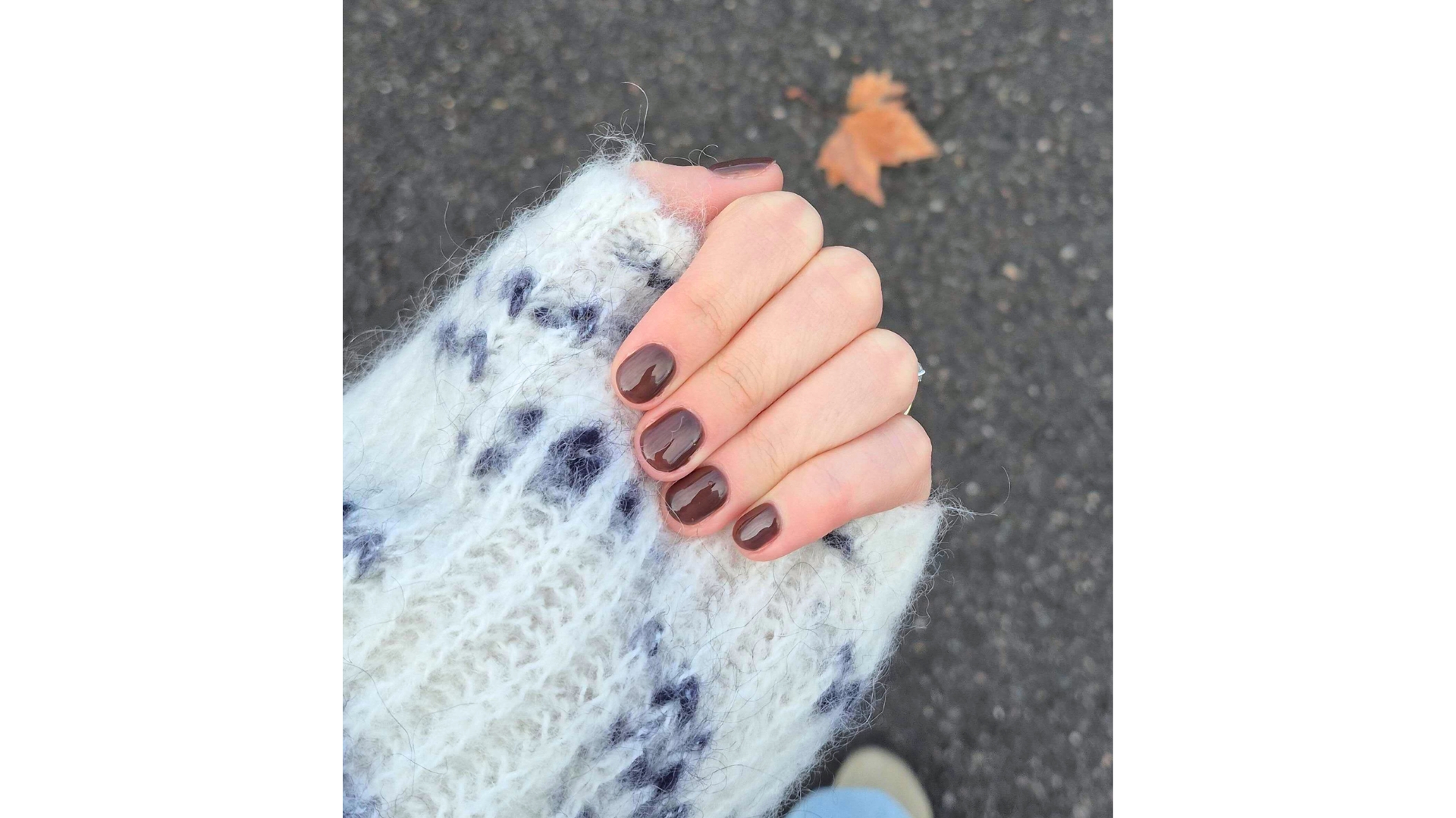 A close-up of Digital Beauty Writer, Naomi Jamieson&#039;s hand, pictured against a white and black knitted jumper and seen with a short chocolate-brown manicure/ above a leafy, concrete background