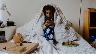 A woman in bed with duvet over her head on phone with pizza boxes next to her