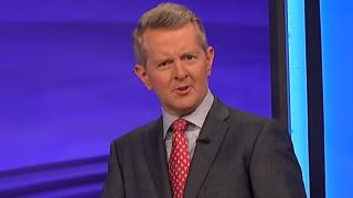 Ken Jennings talking to the camera and wearing a red tie with a white diamond pattern. 