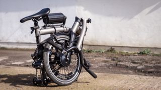 A black and silver folded Brompton T-Line, upgraded with the Cytronex e-bike conversion kit, sits on a rocky surface