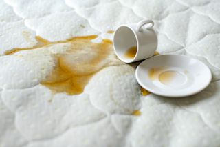 The image shows a cup of coffe tipped on its side with its contents spilling over a white mattress