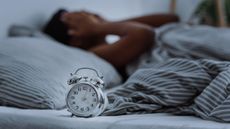 Woman in bed puts hands on face in frustration with alarm clock in foreground