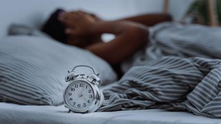 Woman in bed puts hands on face in frustration with alarm clock in foreground