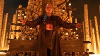 Ralph Fiennes stands in front of a column made of human skulls with his arms open.