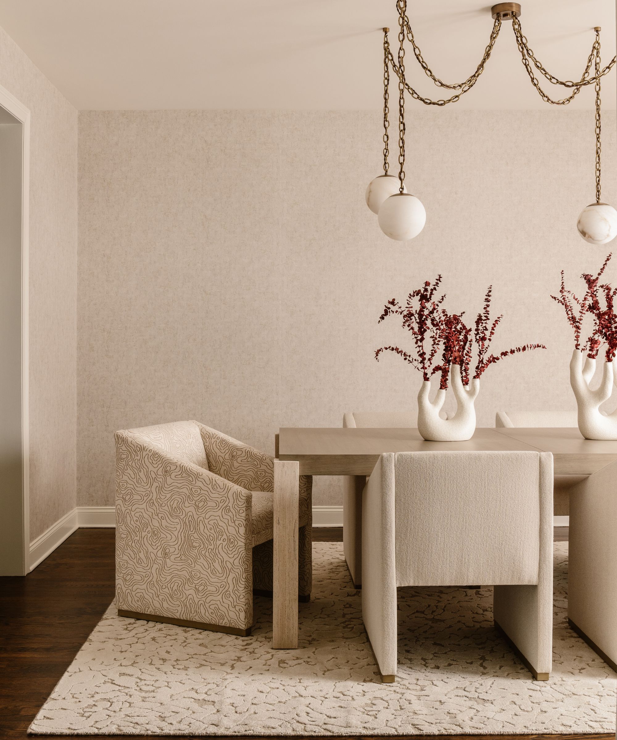 Beige dining room with wooden table and upholstered dining chairs in beige and a beige pattern, a texture beige rug is under the dining table and a statement gold light hangs above