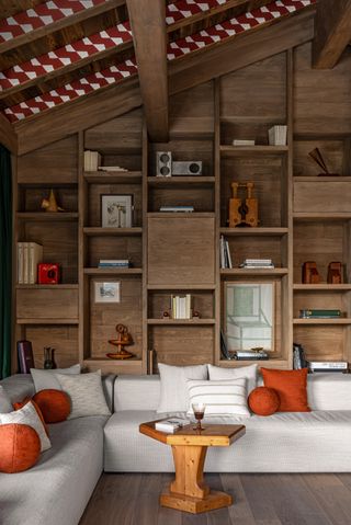 A chalet's attic boasts a floor-to-ceiling bookcase filled with printed matter and sculptural collectibles as well as art and an exaggeratedly voluminous sofa in grey fabric, decorated with orange and striped cushions.