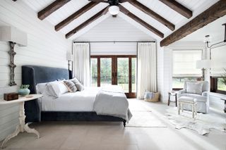large light bedroom with white paneled walls dark beams pale stone floors patio doors and blue bedhead