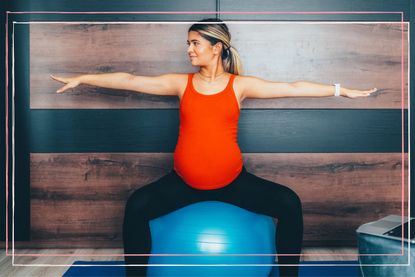 A pregnant woman wearing an orange vest top sits on a Swiss ball doing exercise with her arms out to either side