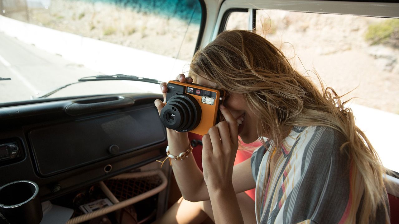 A woman in a campervan taking a picture with an orange Leica Sofort, one of the best instant cameras