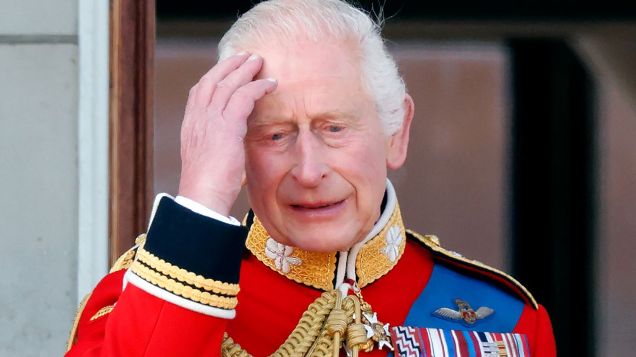 King Charles looks sad and holds his head while wearing a red military uniform