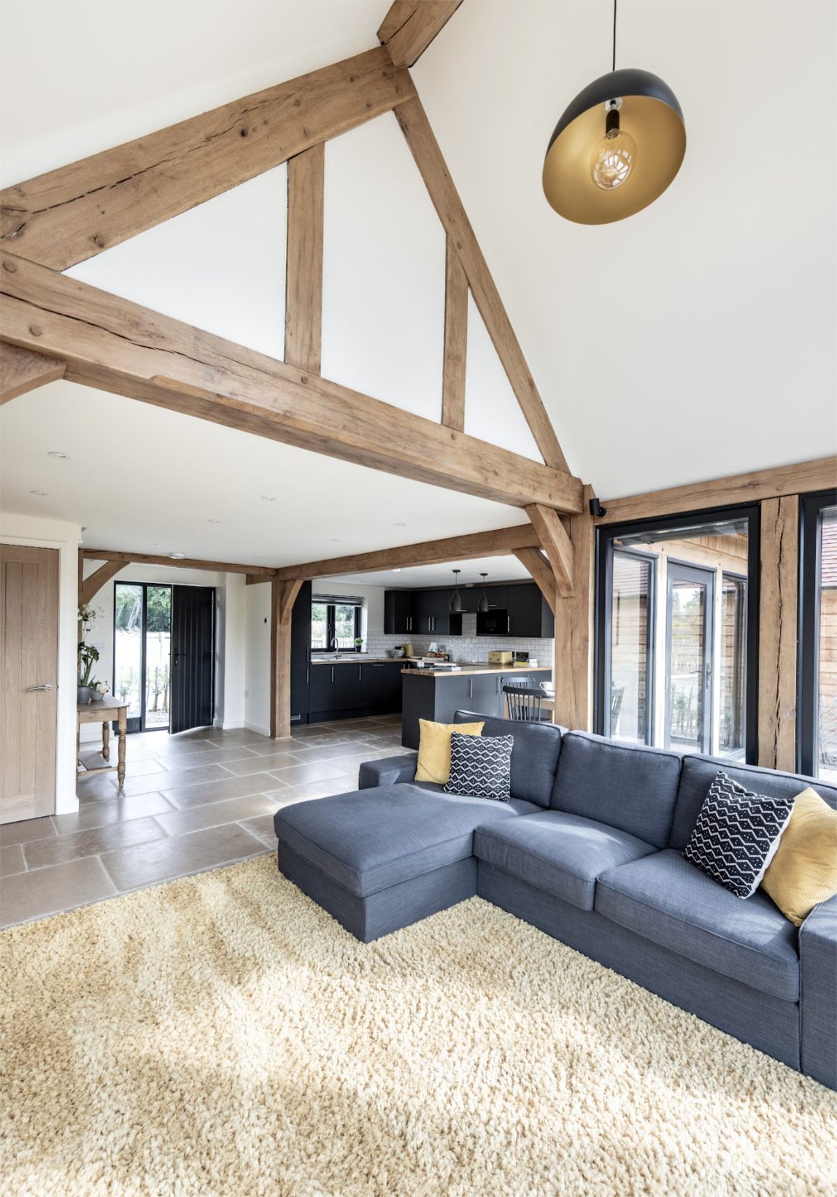 A vaulted open-plan living space with a traditional oak frame aesthetic, a large l-shaped sofa and a large yellow rug